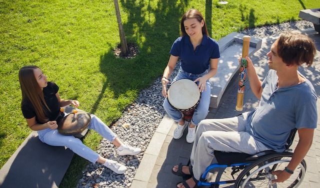 Happy handicapped man on a wheelchair spending time with friends playing live instrumental music outdoors. Concept of social life, friendship, possibilities, inclusion, diversity.